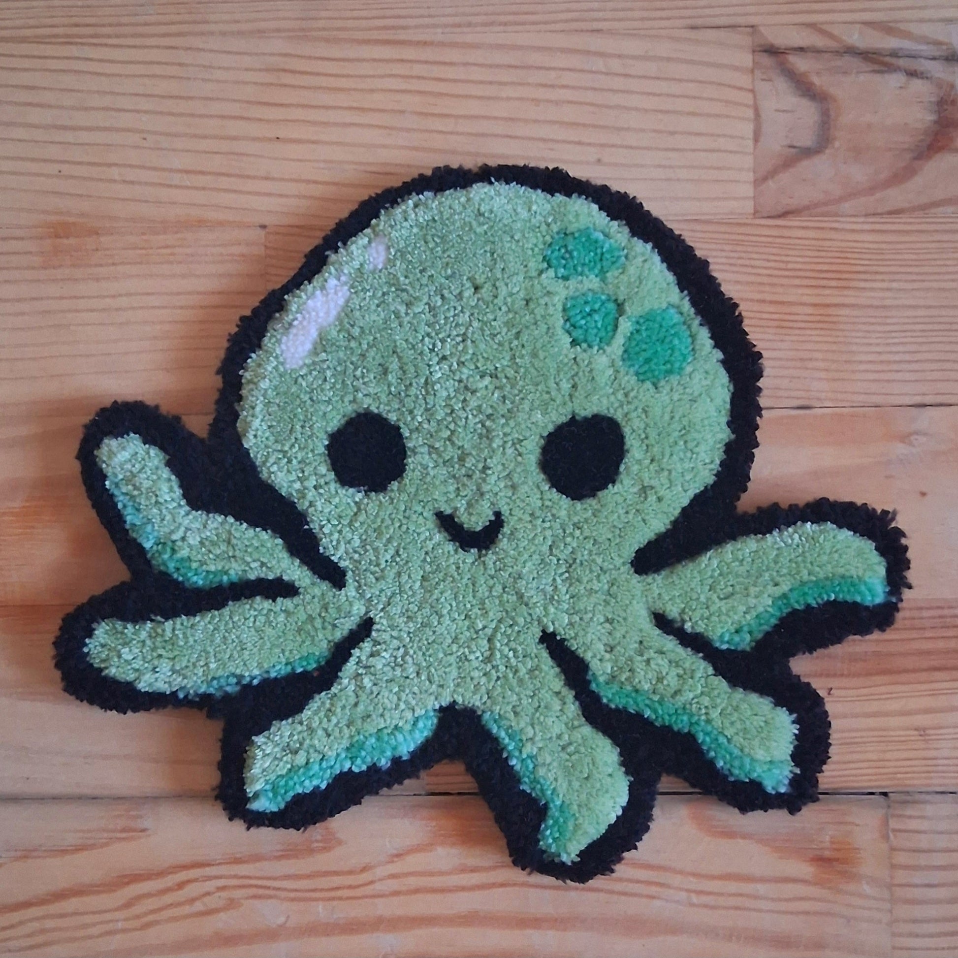Décoration murale représentant un poulpe vert, souriante. Créée par La Balmo Tufting. Laine acrylique verte et noire