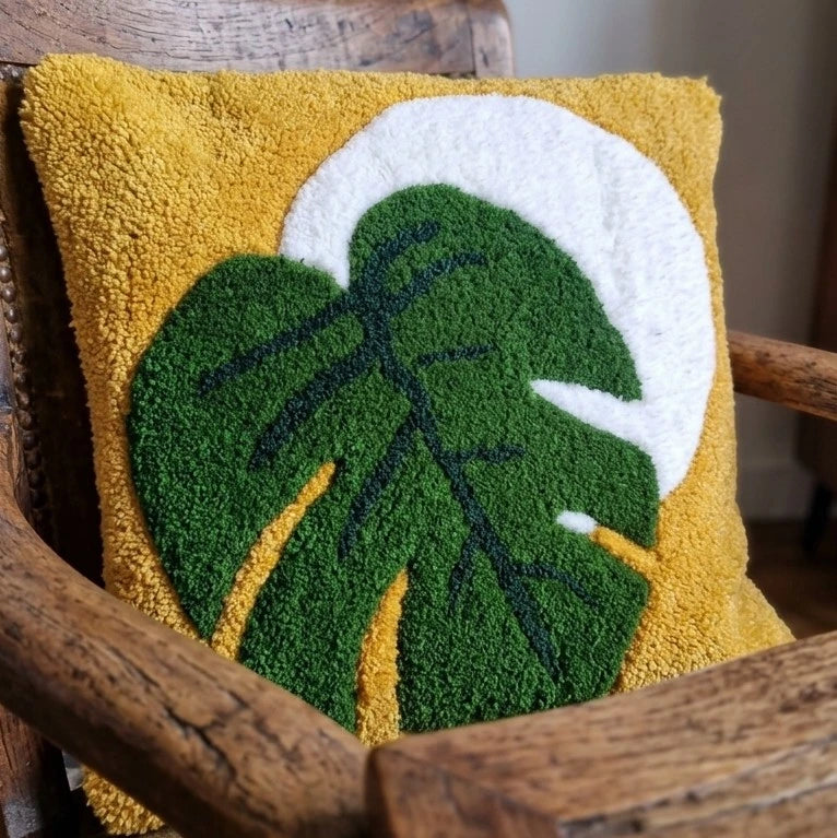 Sur un fauteuil trône un coussin tufté représentant une feuille de monstera sur une lune blanche et un fond jaune. 